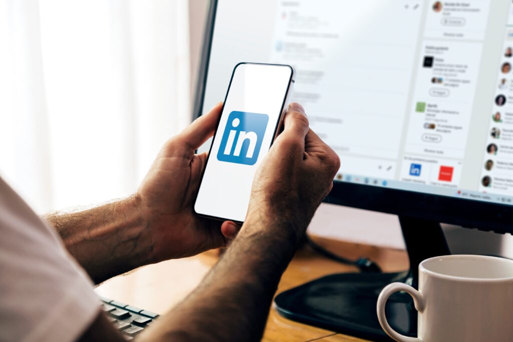 Persona sujetando un móvil con el logo de LinkedIn con un ordeandor en el fondo con la homepage de LinkedIn