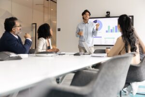 Hombre presentando en una pizara digital ante 2 muejres y 1 hombre